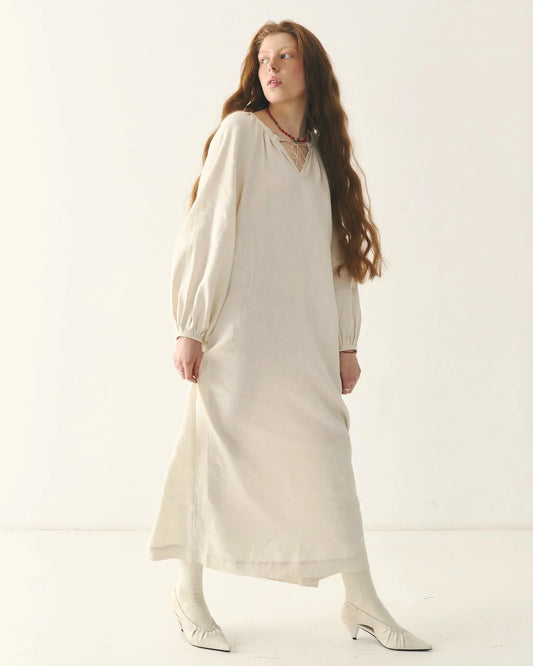 Woman wearing traditional Ukrainian long white linen dress with puff sleeves, standing in a minimalist studio.